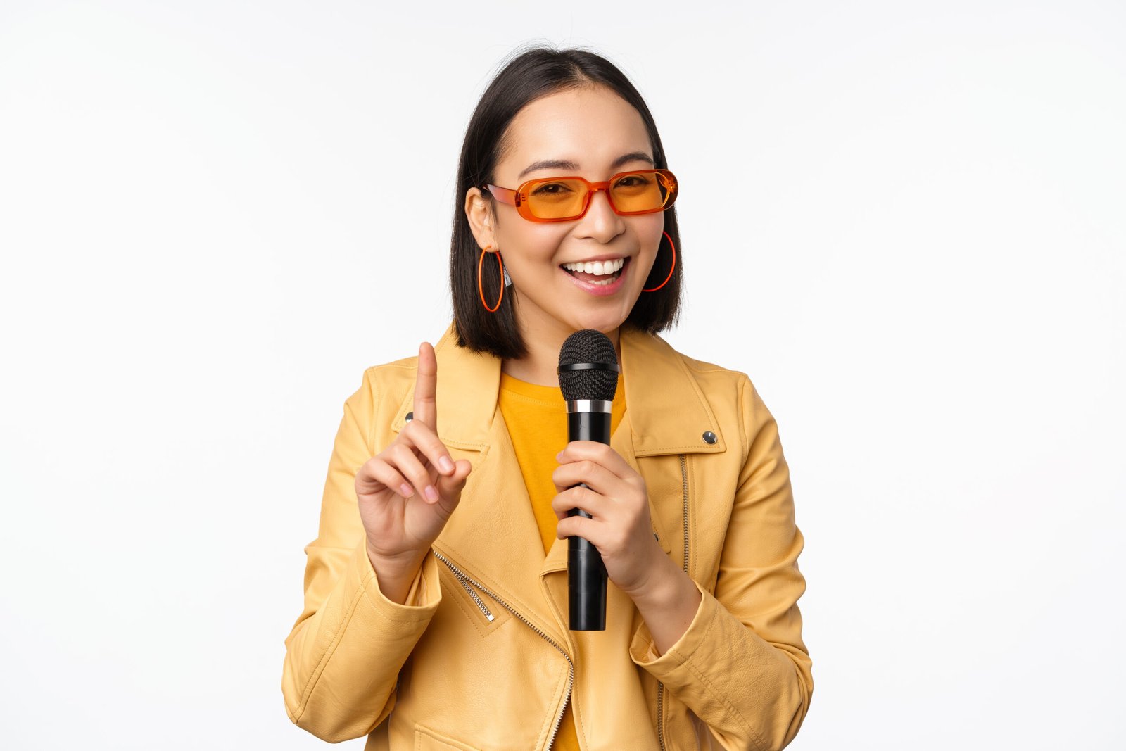 Portrait of beautiful asian woman in sunglasses, stylish girl singing, giving speech with microphone, holding mic and smiling, standing over white background. Copy space