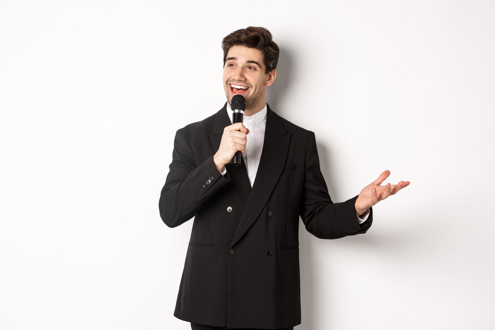 Portrait of handsome man in black suit singing a song, holding microphone and giving speech, standing against white background.